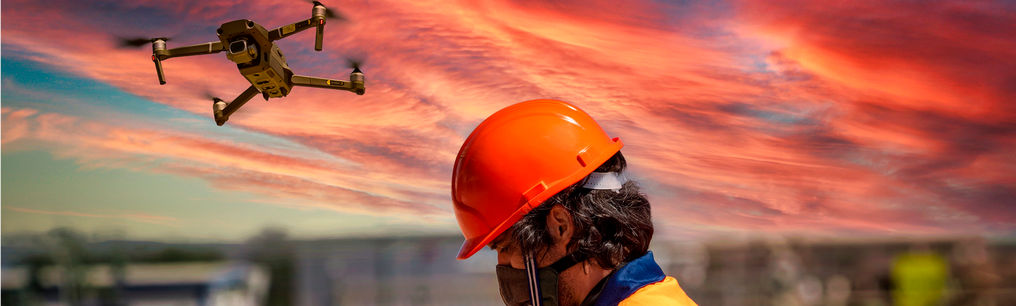 Imatge d'un dron en ple vol al capvespre mentre una persona amb casc de seguretat maneja un control remot